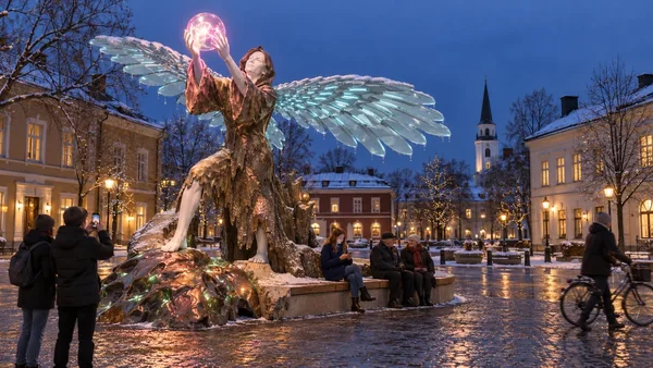 Wi-Fi Angel on a Swedish town square at blue hour: glass wings with cyan light, the glowing sphere held high, people on the bench below