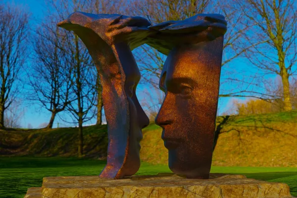 Bronze-look render: two half-faces looking only at each other, joined above by a flowing bridge, on a stone plinth in a park