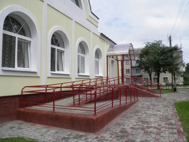 A pale yellow building with arched windows and an entrance ramp in red-brown stamped concrete