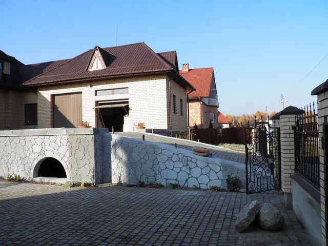 Opěrná zeď se vzorem kamene vedle rampy příjezdové cesty ke garáži, s obloukovým otvorem, na dlážděném předpolí