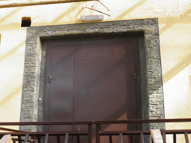 A dark rough-hewn stone surround framing a metal door on a yellow building